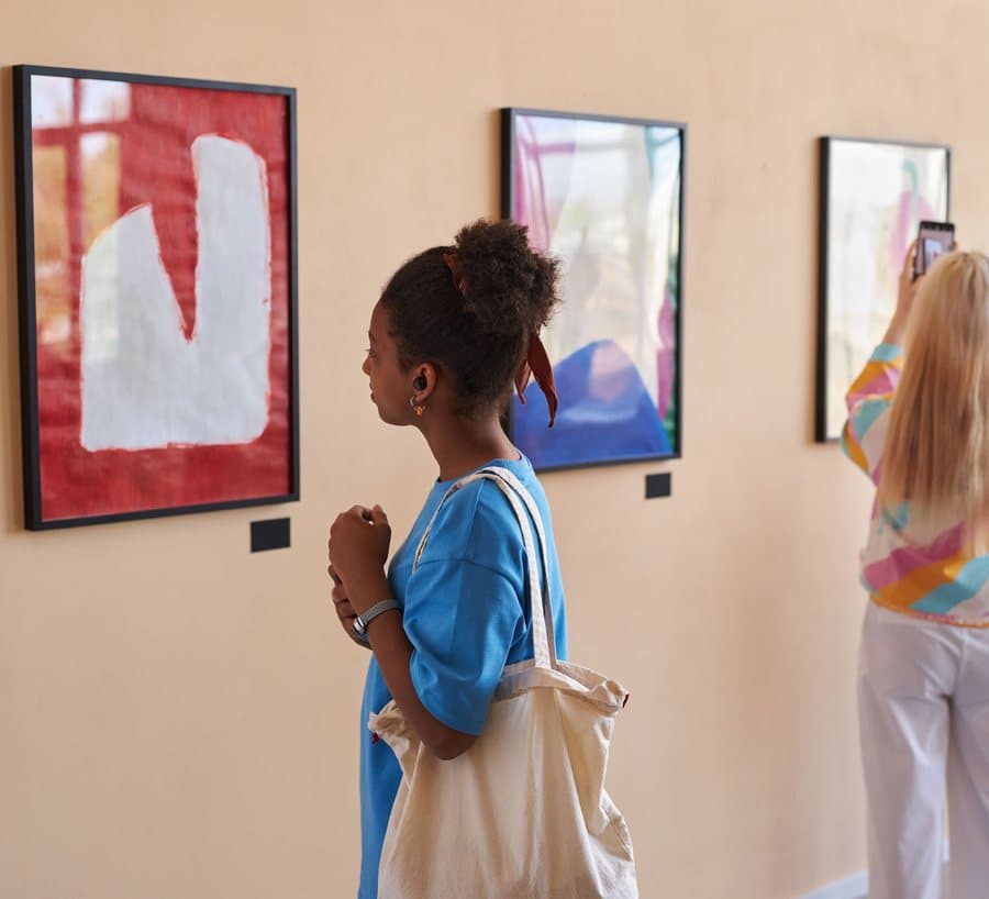 Young visitors exploring an art gallery, one photographing a painting with her phone.